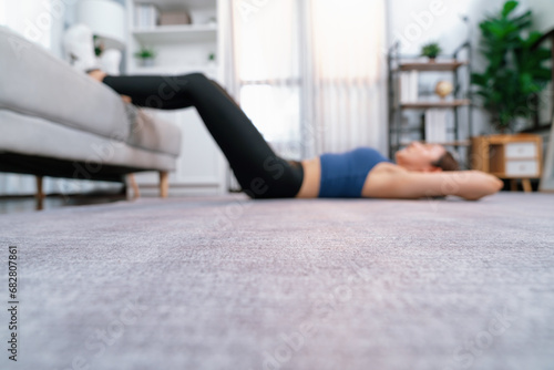 Blurred background of young attractive asian woman in sportswear lies on house floor and doing sit-ups on the crouch, aiding targeting on abs muscle for effective home exercise routine. Vigorous