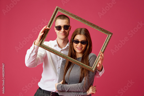 Young couple, man and woman in sunglasses posing with picture frame against pink studio background. Bonding. Concept of love, relationship, Valentine's Day, emotions, lifestyle