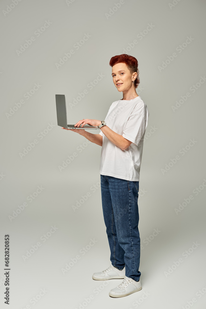 redhead queer freelancer in white t-shirt standing with laptop and looking at camera on grey redhead queer freelancer in white t-shirt standing with laptop and looking at camera on grey