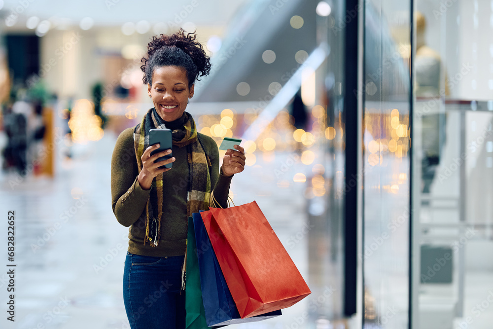 Wall Mural Happy black woman using smart phone and credit card while checking her bank account after shopping in mall