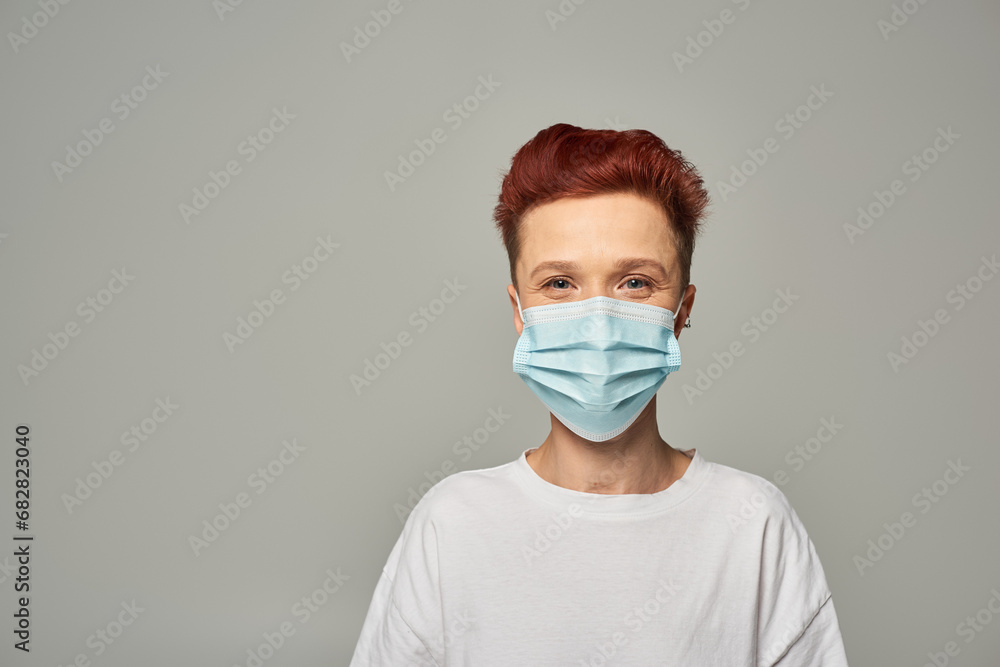 portrait of redhead queer person with happy gaze wearing medical mask and looking at camera on grey portrait of redhead queer person with happy gaze wearing medical mask and looking at camera on grey