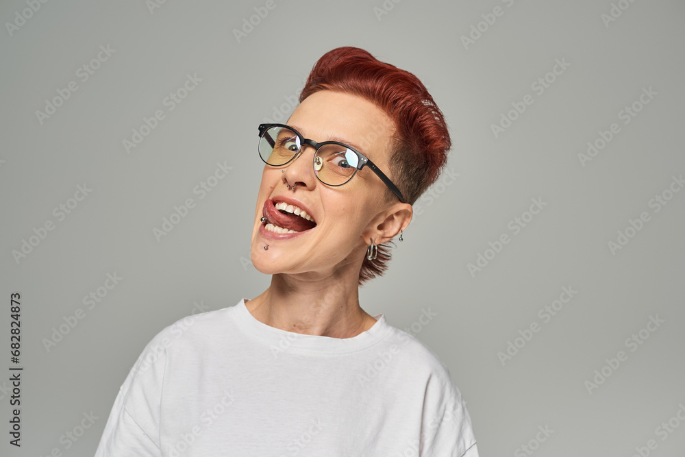 funny and cheerful non-binary person in white t-shirt and eyeglasses sticking out tongue on grey
