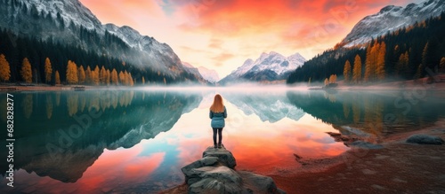 Woman in red jacket is standing on the coast of mountain lake at sunrise. AI generated image