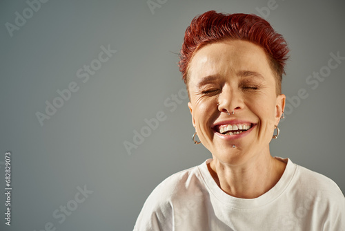 portrait of redhead bigender person with facial piercing laughing with closed eyes on grey backdrop