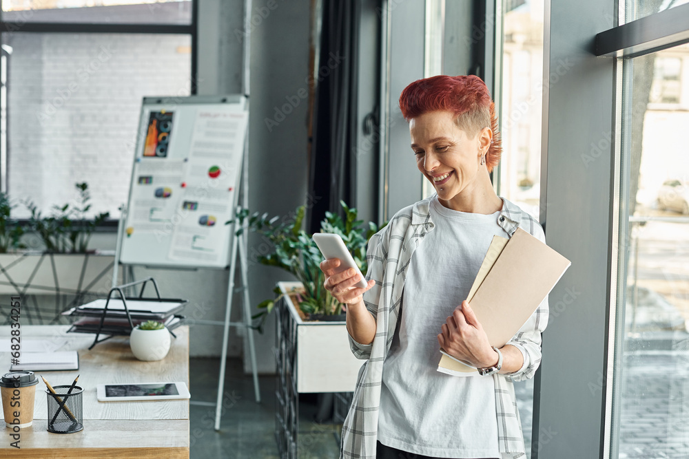 happy queer person in casual attire standing with documents in office and messaging on smartphone happy queer person in casual attire standing with documents in office and messaging on smartphone