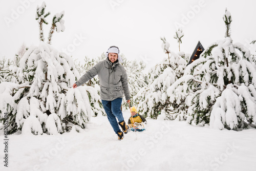 Father pulling little son on winter day. Children are rolling down hill on sledge in forest. Happy funny child with dad ride sled on snowy road in mountains. Family walks during snowfall in park. photo