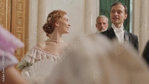Medium shot of diverse pairs of beautiful debutantes and their male partners wearing adorable Regency outfits strolling along luxury ballroom before dancing photo
