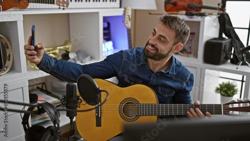 Young, handsome hispanic man thoughfully strumming a melody on his classical guitar, taking a vibrant selfie image with his smartphone in the pulsating heart of the music studio. photo