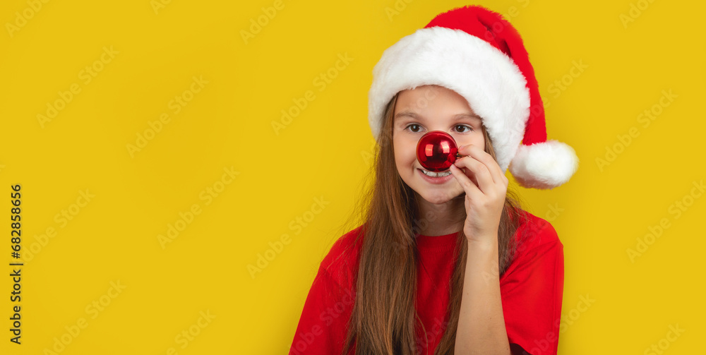 Holiday Fun. Cheerful Little Girl Wearing Santa hat, Playful Kid Keeping Xmas Tree Ball Near Nose on Yellow Background. Copy Space Holiday Fun. Cheerful Little Girl Wearing Santa hat, Playful Kid Keeping Xmas Tree Ball Near Nose on Yellow Background. Copy Space