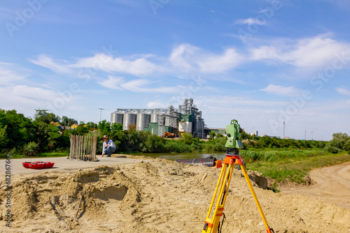 Geodesist device on tripod at building site over water are big grain silos in industrial landscape