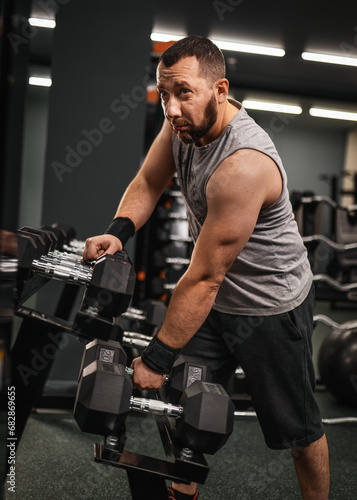 A bearded male bodybuilder takes a dumbbell in his hand and demonstrates the triceps. Work on your body in the gym.