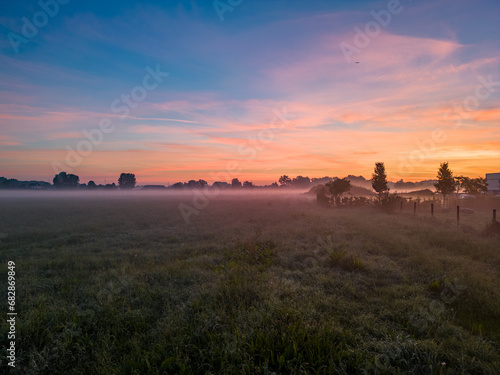 The photograph exudes the ethereal charm of early morning on a meadow, with the first light of daybreak painting the sky in hues of pink, orange, and blue. A delicate mist hovers above the ground