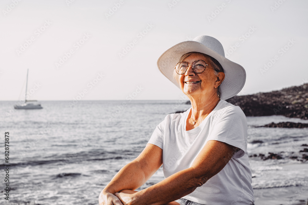 Carefree satisfied senior woman with white hat looking at new day from sea beach at sunrise enjoying freedom, vacation or retirement concept