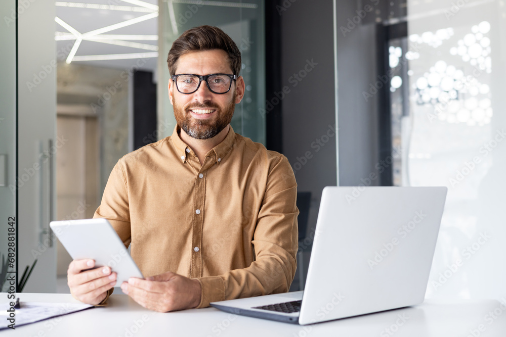 Portrait of a young businessman working in an office center, reading at a table with a laptop and holding a tablet in his hands, smiling and looking at the camera Portrait of a young businessman working in an office center, reading at a table with a laptop and holding a tablet in his hands, smiling and looking at the camera