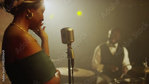 Waist up of modern African American jazz dup giving concert in fancy jazz club in evening. Young woman singing in mic while man playing drums in background on smoke filled stage