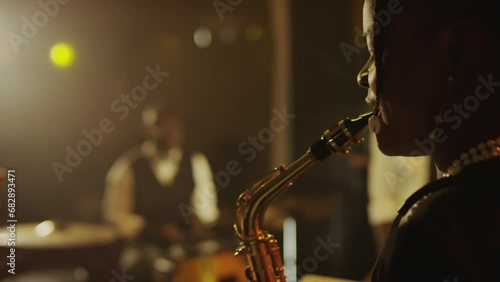 Side view medium closeup of young Black female saxophonist playing instrument while performing on stage with jazz band on stage in fancy club photo