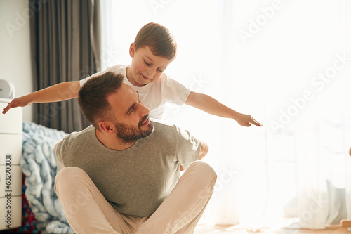 On the father shoulders. Man is playing with his little son in the domestic room