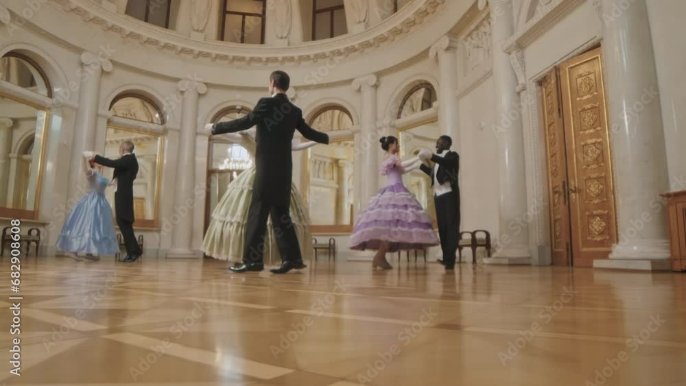 Slowmo long shot of beautiful women in poof dresses and their male companions ball dancing on parquet floor in spacious classic ballroom with ornamental walls and windows Slowmo long shot of beautiful women in poof dresses and their male companions ball dancing on parquet floor in spacious classic ballroom with ornamental walls and windows