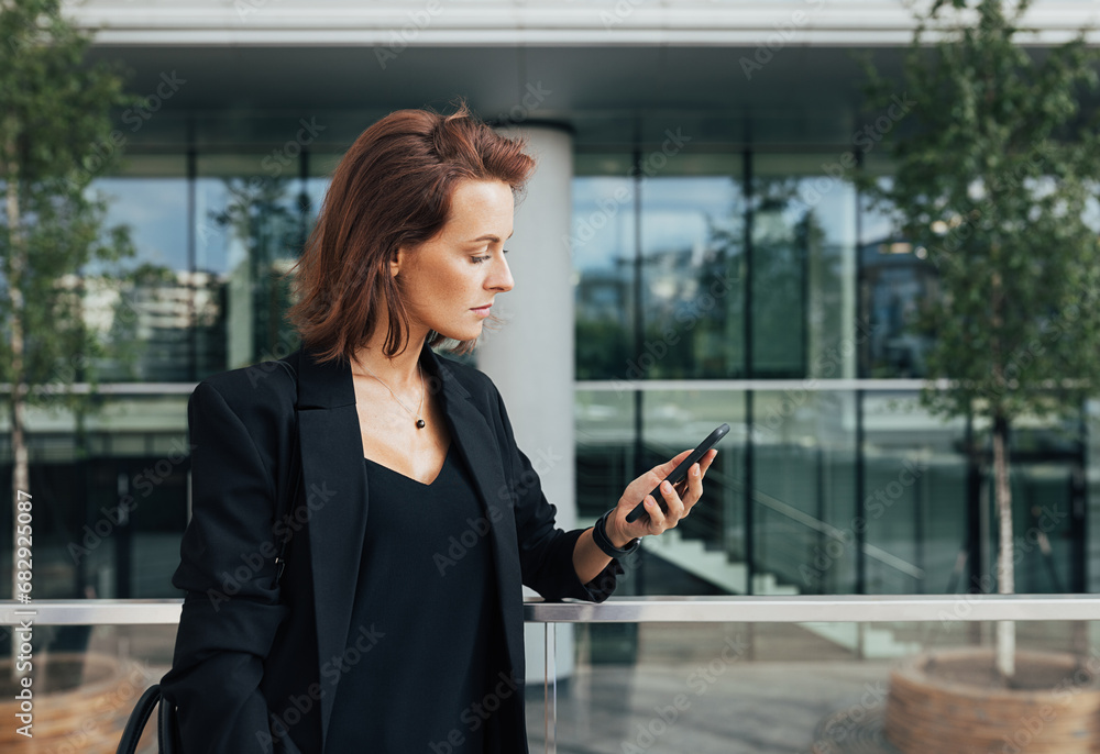Fototapeta premium Side view of a confident businesswoman with ginger hair looking at her smartphone against an office building