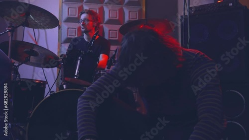 A singer is emotionally singing a song into a microphone, dramatically sitting on the floor in a music studio next to speakers and a drum kit on which a drummer is playing. photo