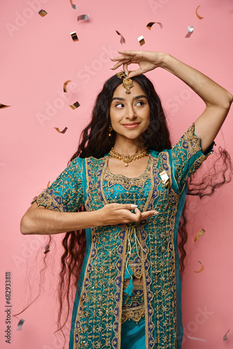 jolly attractive indian woman gesturing and looking away under confetti rain on pink backdrop
