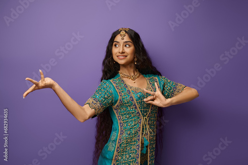 jolly indian woman in traditional clothing gesturing while dancing and looking away, purple backdrop