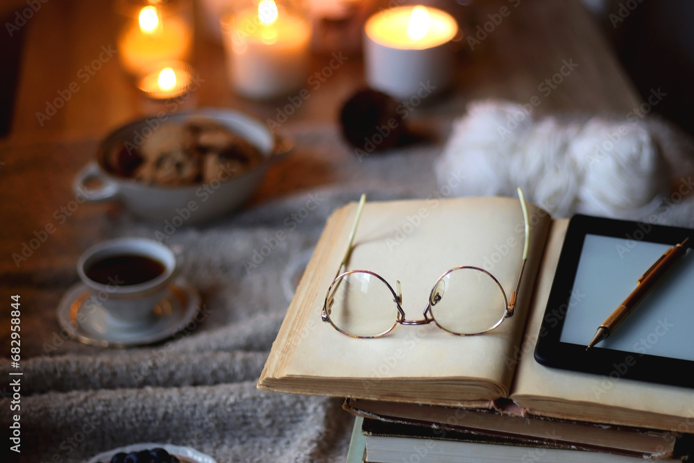 Fototapeta premium Bowl of cookies, cup of tea or coffee, plate of blueberries, dry oranges, wool yarn, pine cones, books, reading glasses, tablet, pen, soft blanket and lit candles on the table. Winter hygge at home.