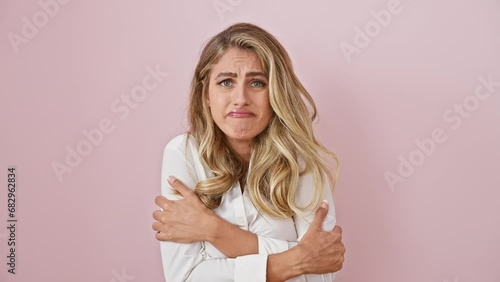 Heartbroken young blonde woman in shirt shivers, sad expression on her face from winter's cold shock, standing alone against pink background photo