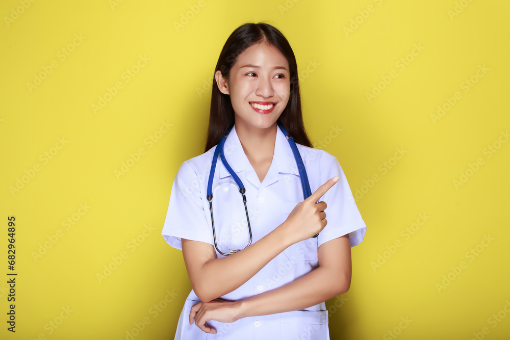 Portrait of a beautiful young woman in a yellow background, Friendly beautiful woman wearing a doctor's uniform and pointing while wearing a stethoscope. Portrait of a beautiful young woman in a yellow background, Friendly beautiful woman wearing a doctor's uniform and pointing while wearing a stethoscope.