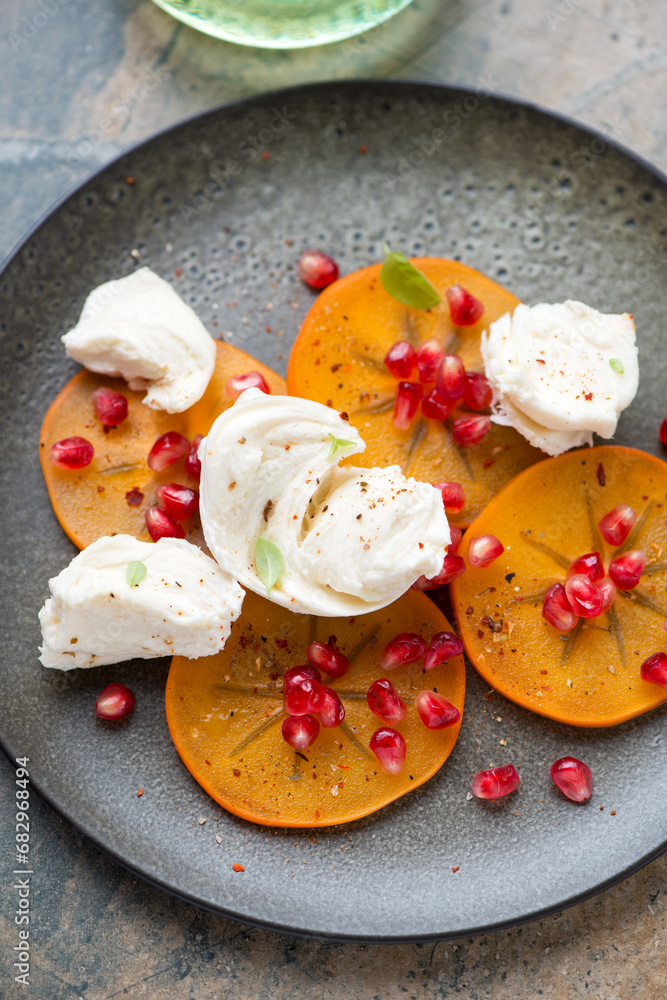 Obraz premium Olive-colored plate with persimmon slices, torn mozzarella and pomegranate seeds, vertical shot, middle closeup