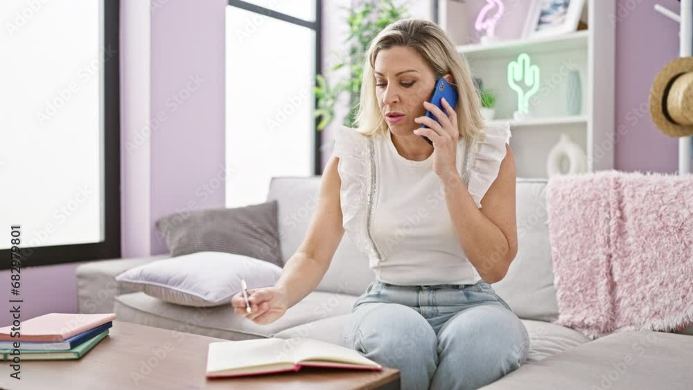 Young blonde woman talking on smartphone taking notes sitting on sofa at home Young blonde woman talking on smartphone taking notes sitting on sofa at home