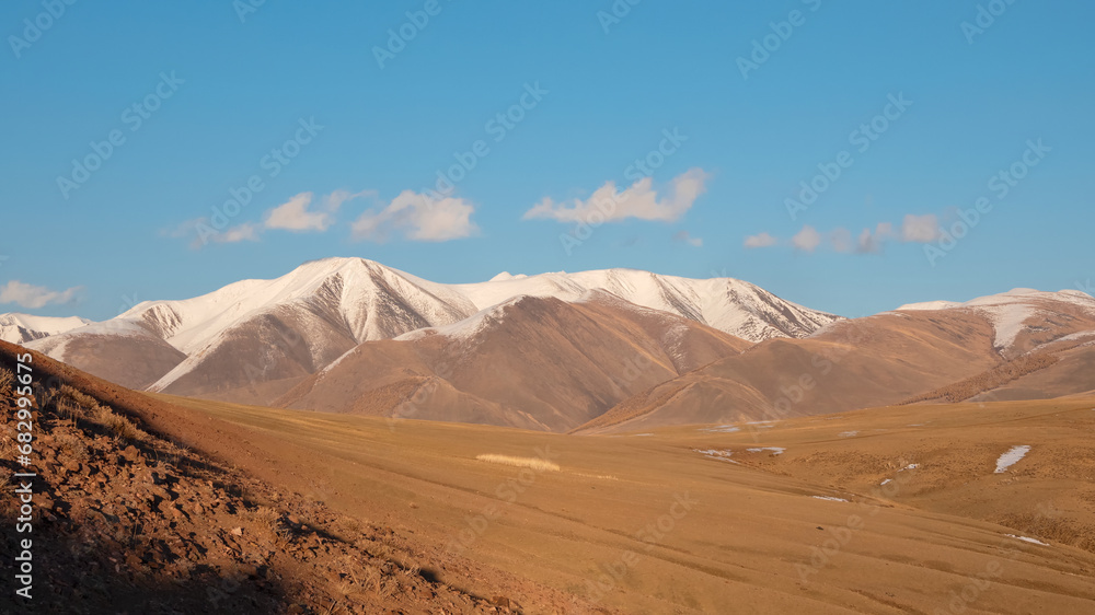 Wide panoramic view of bright autumn mountain steppe landscape. Awesome alpine view from pass to mountain valley in sunlight and great mountain silhouettes on horizon with white clouds on blue sky. Wide panoramic view of bright autumn mountain steppe landscape. Awesome alpine view from pass to mountain valley in sunlight and great mountain silhouettes on horizon with white clouds on blue sky.