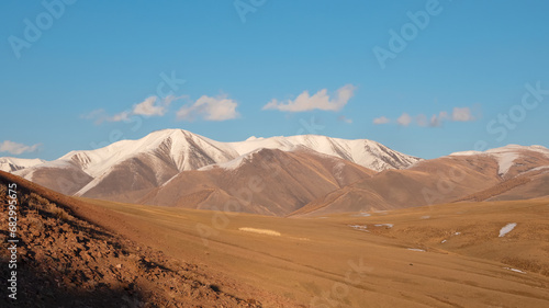 Wide panoramic view of bright autumn mountain steppe landscape. Awesome alpine view from pass to mountain valley in sunlight and great mountain silhouettes on horizon with white clouds on blue sky.