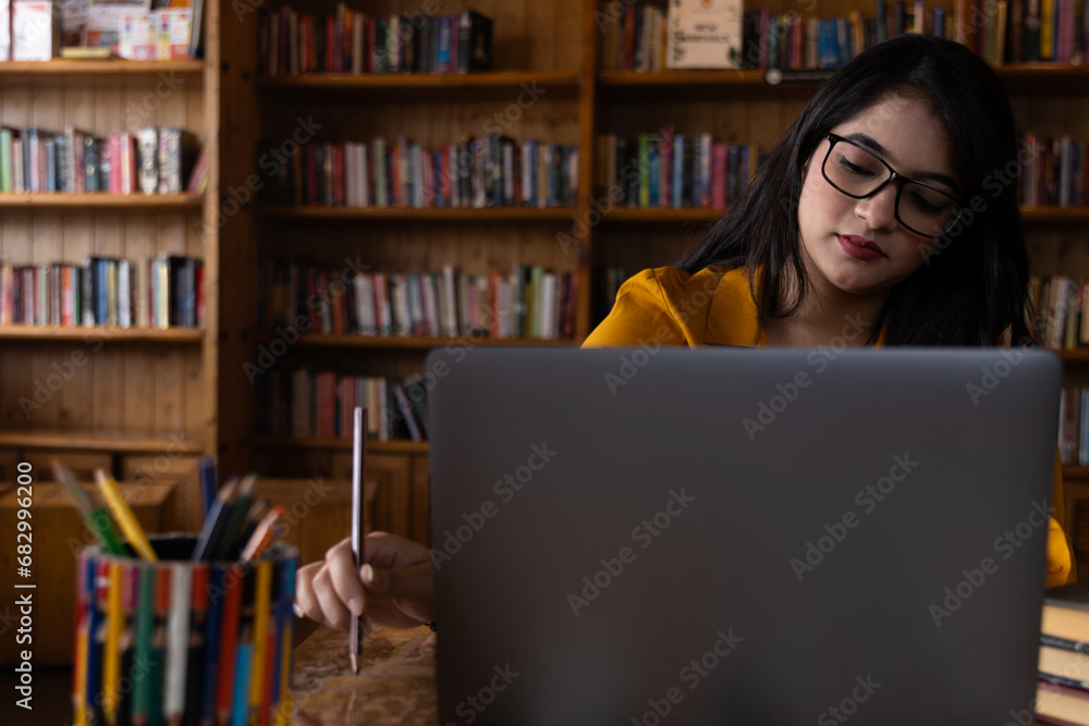 A young girl student wearing glasses or spectacles and studying seriously in school or collage library or working in corporate office using laptop searching for information on internet or books A young girl student wearing glasses or spectacles and studying seriously in school or collage library or working in corporate office using laptop searching for information on internet or books