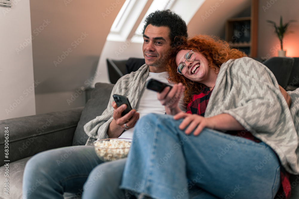 Interracial couple sitting on couch, snuggled in blankets, eating popcorn and scrolling through movies on television while talking