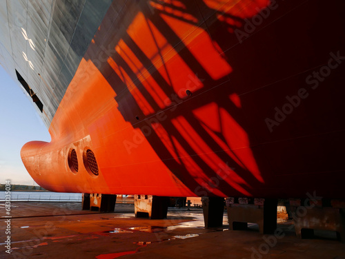 Ship in dry dock on keel blocks. Bow thrusters and anchor. Red and black hull with sunset shadows. 