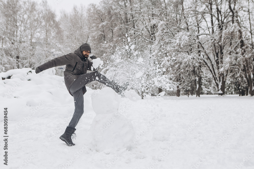 Young man having fun destroying breaking snowman in winter snowy season in city park covered hill, holiday vacation weekend, enjoying spending time together
