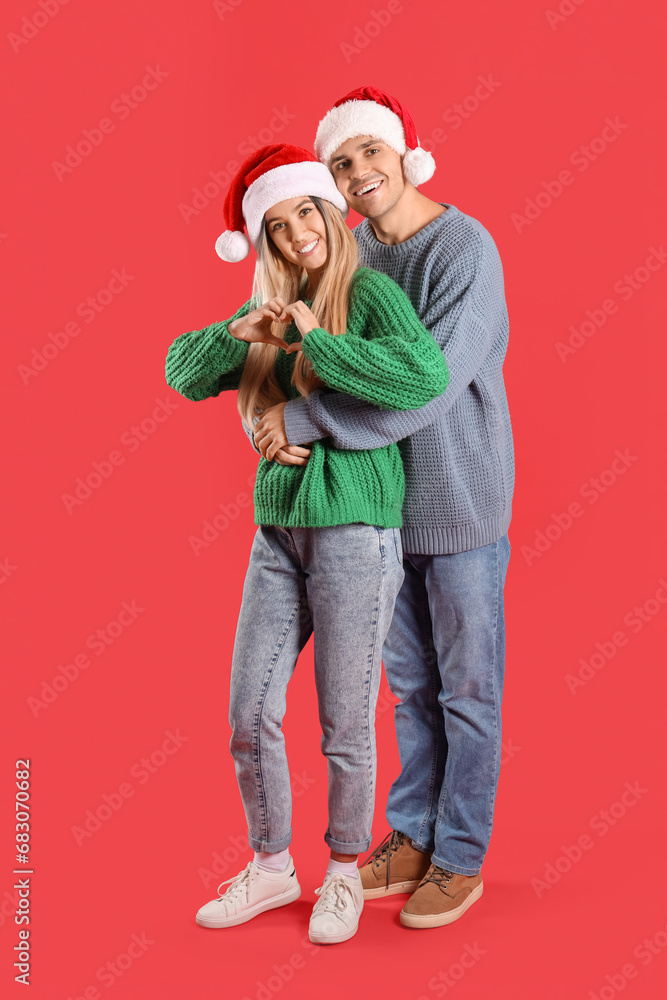 Happy young couple in Santa hats making heart gesture on red background Happy young couple in Santa hats making heart gesture on red background
