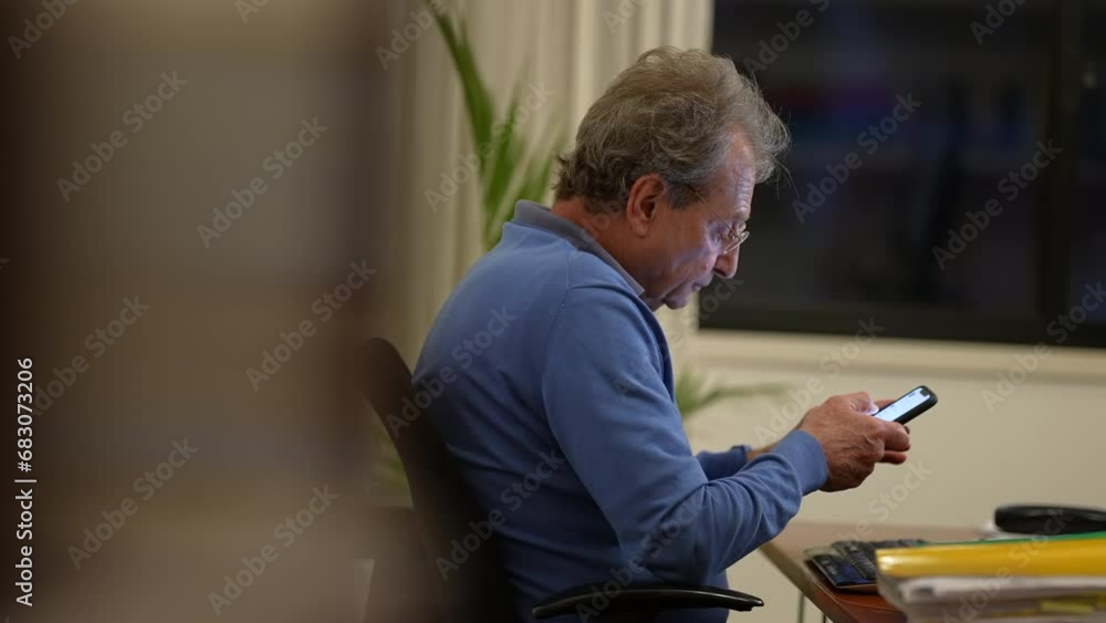 One candid older entrepreneur reading message on cellphone device seated at office at night. 70s male mature business man working One candid older entrepreneur reading message on cellphone device seated at office at night. 70s male mature business man working