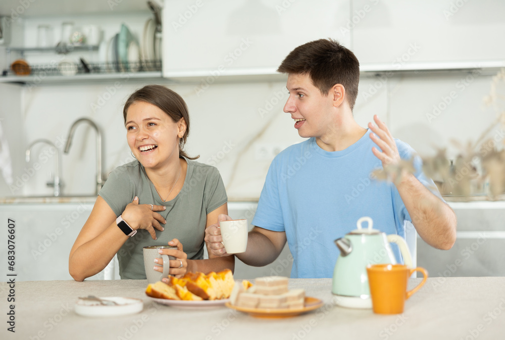 Portrait of cheerful young couple, girl and guy, drinking tea, talking friendly and laughing at table in cozy home kitchen ..