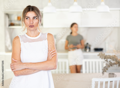 Offended mother standing in the kitchen folding her arms with her back to upset young daughter standing at the kitchen-cupboard
