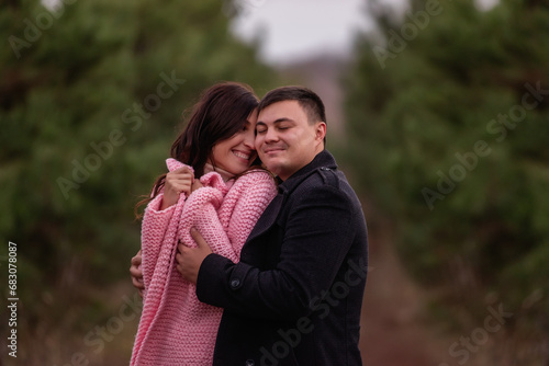 Close-up portrait of happy couple hugging in green coniferous forest. Young man tactilely wrapped black coat around curly-haired woman in pink sweater. Winter engagement, autumn wedding. Marriage