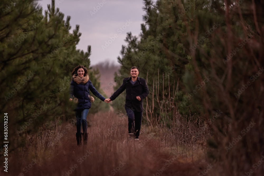 Happy couple running through the forest holding hands. Man in black coat and a young woman in a knitted sweater are fooling around in a pine park, playing tag. Winter date in nature. valentines day
