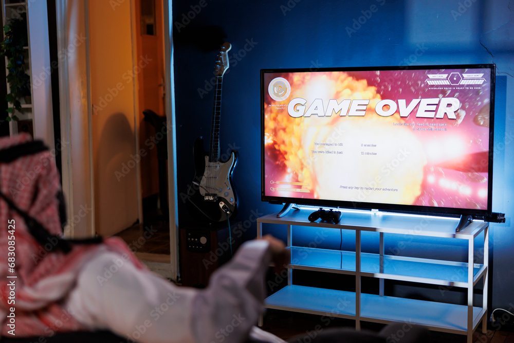 Islamic guy defeated in an online video game from the comfort of his home. Lit by the glow of the screen, he grasps a controller and uses a wireless joystick for immersive gameplay.