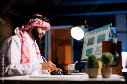 A Muslim businessman in traditional attire uses a pen to jot down notes from his computer screen. The image portrays a diligent Arab guy conducting a research study with a desktop pc and notepad. photo