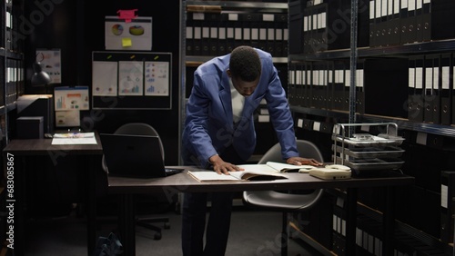 African american private investigator standing and cross-checking clues and evidence with laptop. Black policeman with criminology knowledge examines forensic files and classified research data.
