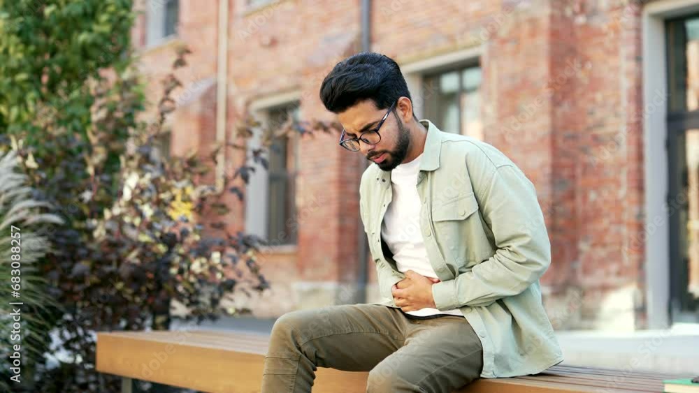 A student is suffering from a stomach ache sitting on a bench in the campus space near university building. Upset male has food poisoning, gastritis, ulcer, constipation, digestive problems or cramps A student is suffering from a stomach ache sitting on a bench in the campus space near university building. Upset male has food poisoning, gastritis, ulcer, constipation, digestive problems or cramps