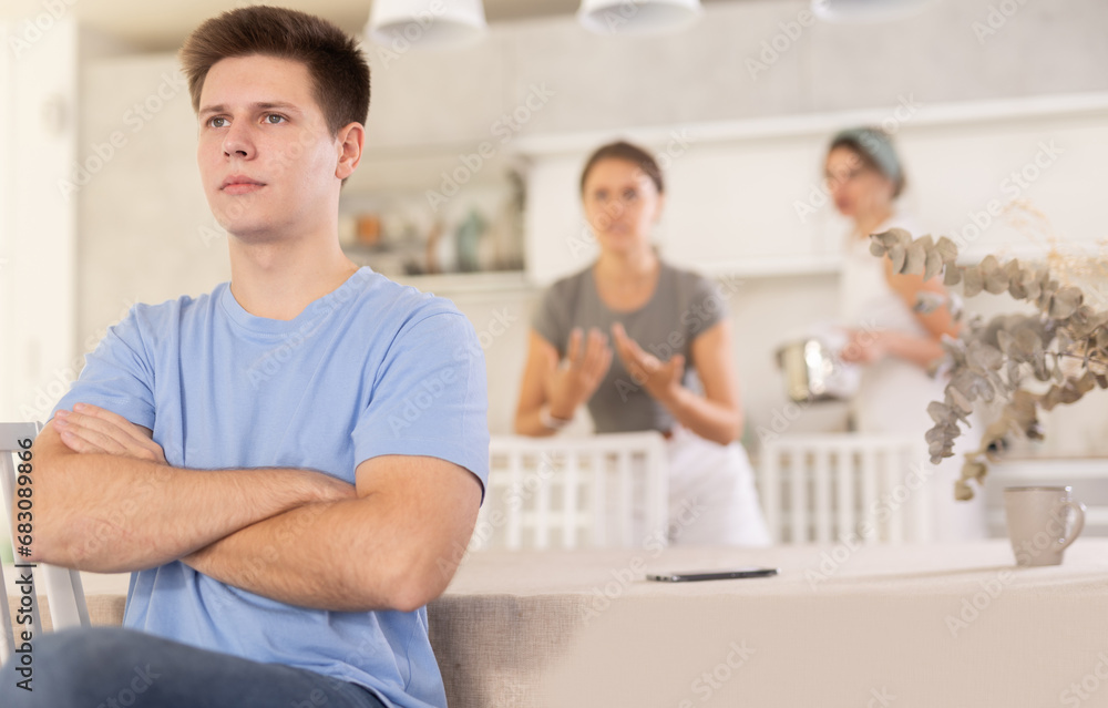 Fototapeta premium Woman mother and young girl during family quarrel aggressively argue with adult son, prove his wrong. Unconcerned guy sits with back to mother and sister, ignores claims of screaming mother