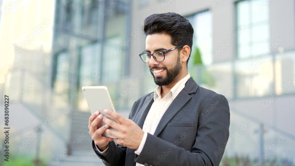 Smiling young businessman in formal suit is using a tablet while standing on street near an office building. Happy entrepreneur reading checking email, chatting, texting messages or shopping online Smiling young businessman in formal suit is using a tablet while standing on street near an office building. Happy entrepreneur reading checking email, chatting, texting messages or shopping online