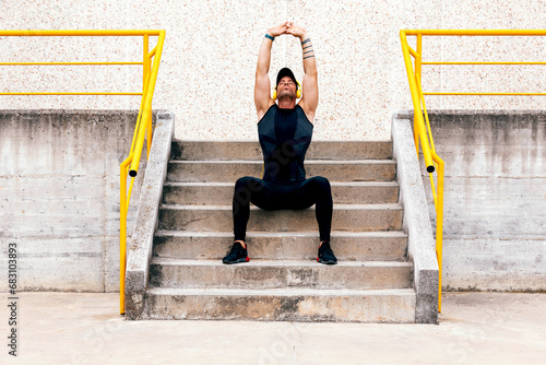 Full length of muscular sportsman with eyes closed outstretching arms while listening music over yellow headphones while sitting on concrete steps in urban city photo